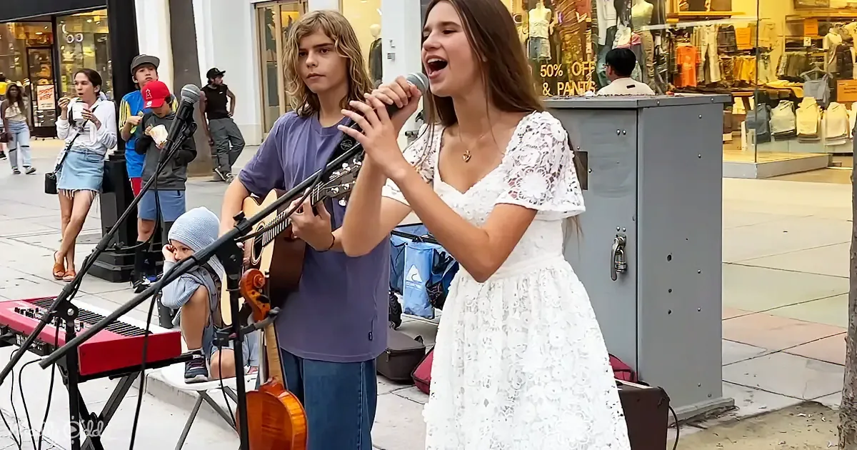 A young woman in a white dress sings into a microphone while a boy plays guitar in a street performance.