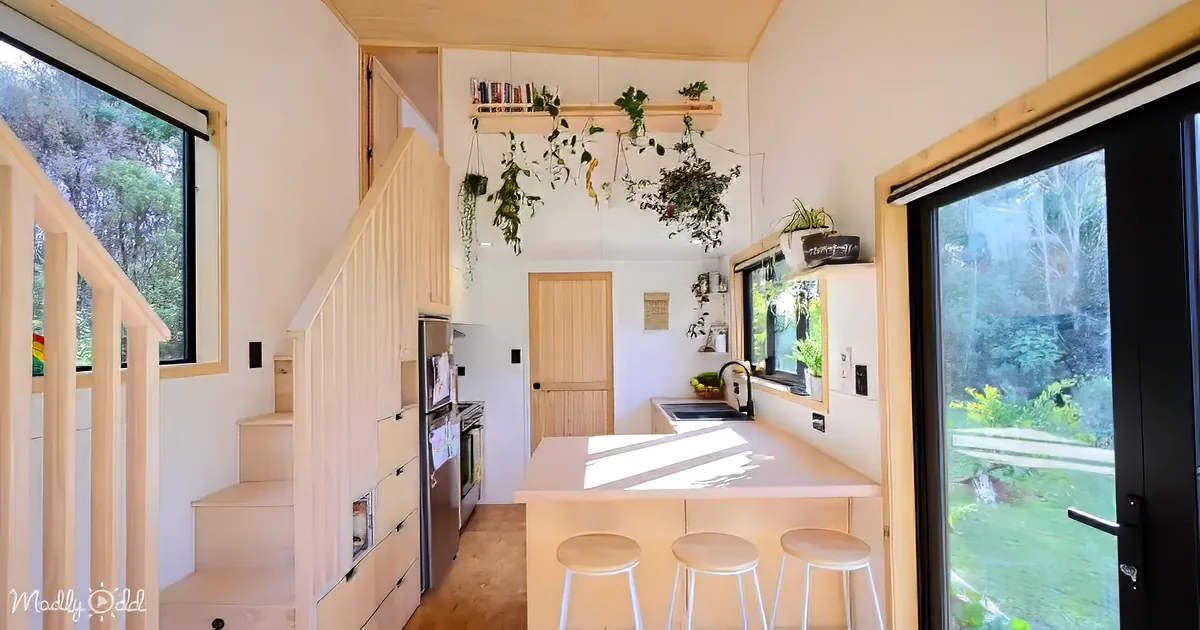 A cozy, compact kitchen with a central island, stools, hanging plants, and a view of the outdoors through a sliding door.