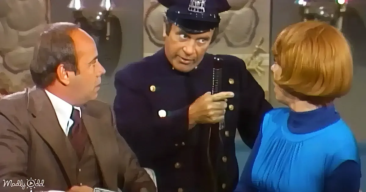 A man in a police uniform holds a microphone, speaking to two others, one in a suit and the other in a blue dress.