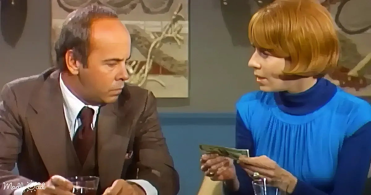 A man in a suit sits at a table, looking down, while a woman in a blue dress holds a small object and speaks.