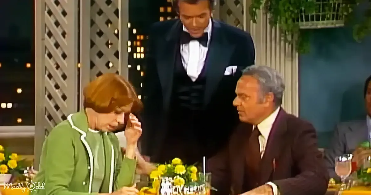 A woman in a green jacket sits at a table with a man in a suit, while a waiter in a tuxedo stands behind them.