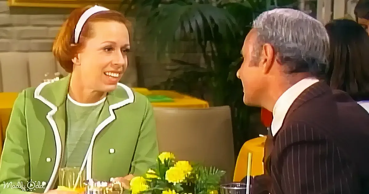 A woman in a green jacket smiles at a man in a pinstripe suit at a table with yellow flowers.