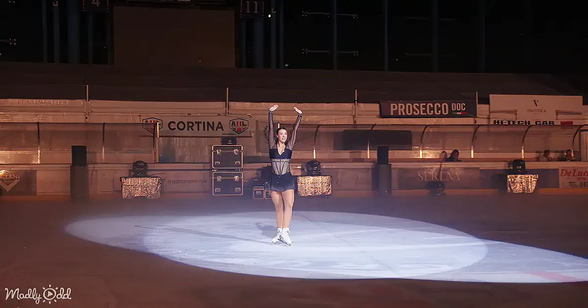 A figure skater in a black outfit stands on ice with arms raised, spotlighting her.