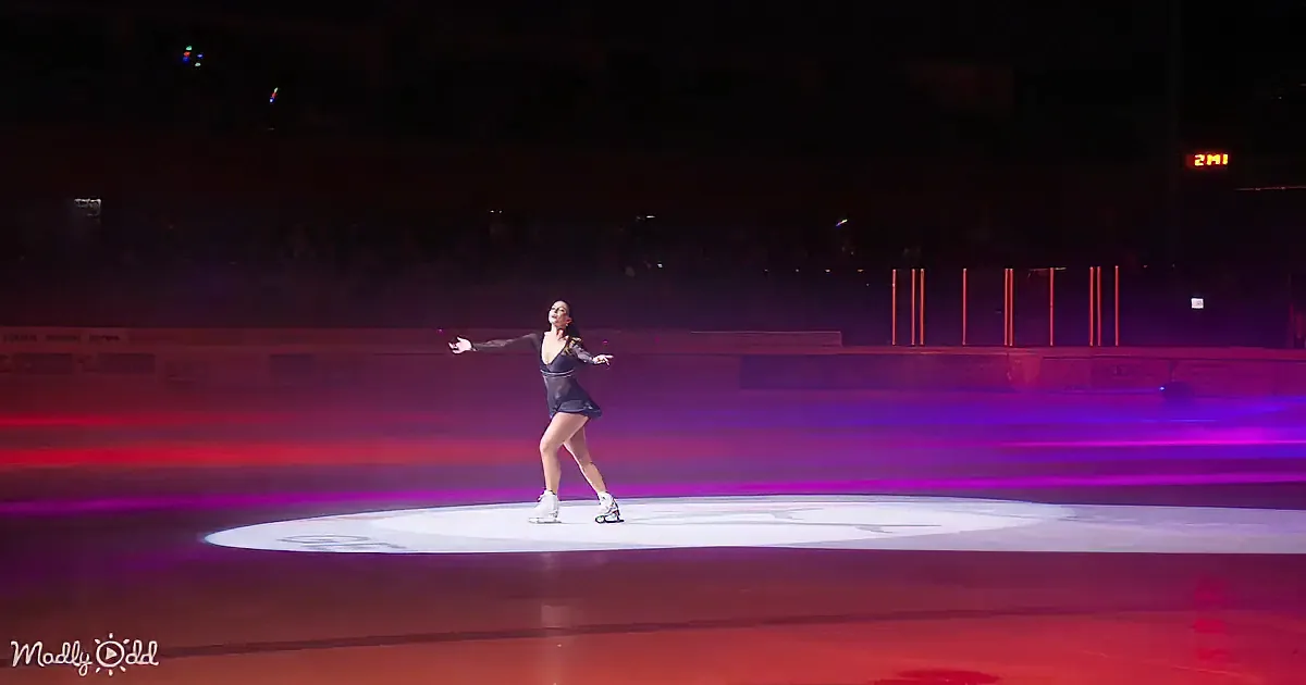 A figure skater in a dark outfit glides gracefully on an ice rink under vibrant purple and red lights.