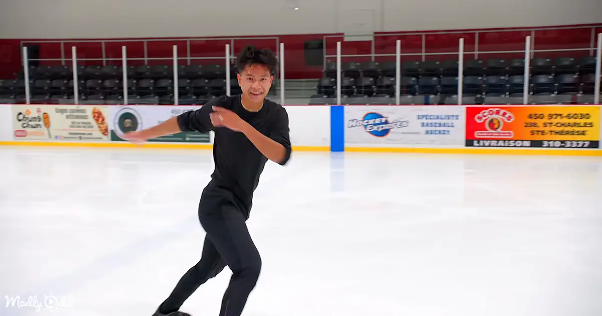 A person in black attire skates on an ice rink, arms outstretched, with advertisements and empty seats in the background.