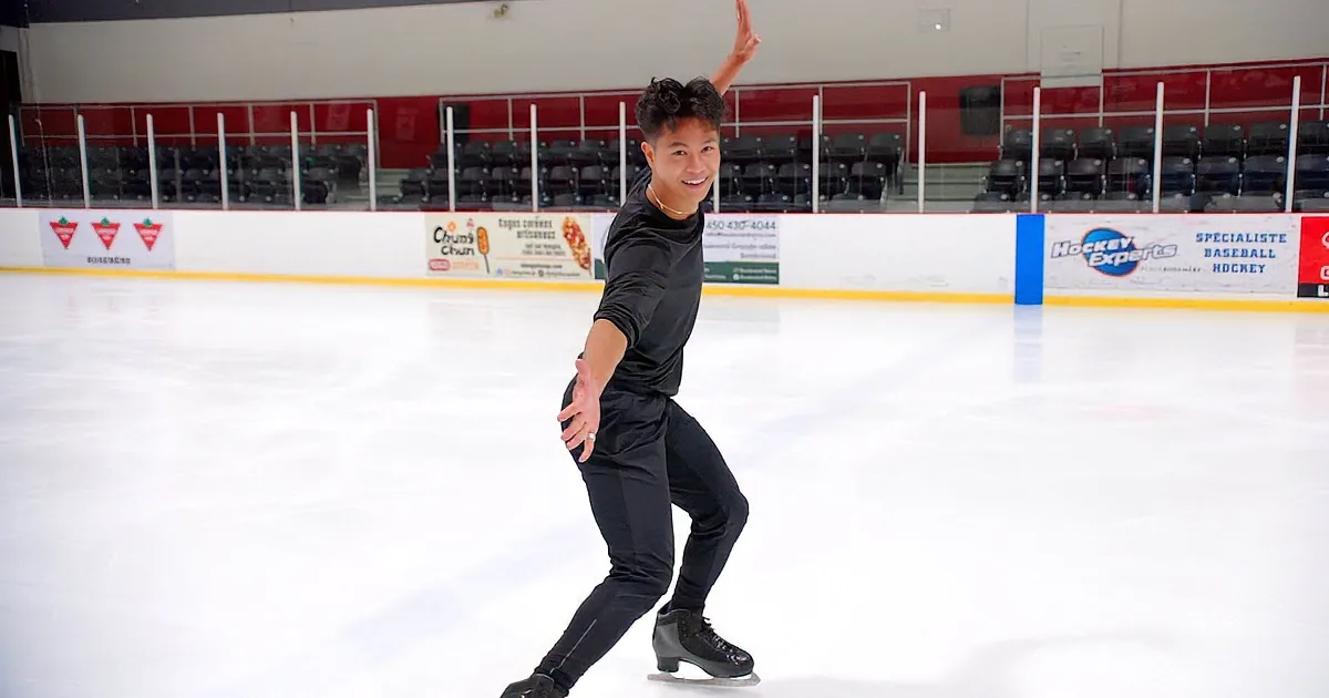A person in black attire skates on an ice rink, arms outstretched, with advertisements on the boards in the background.