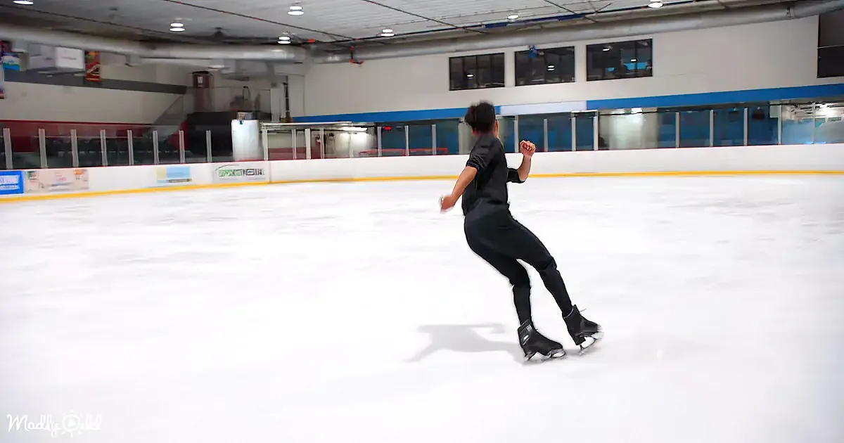 A person in black attire skates on an indoor ice rink, surrounded by walls and advertisements.