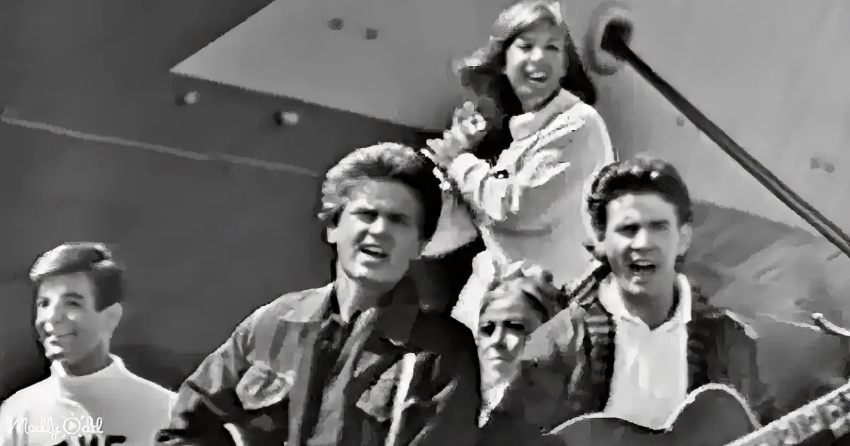 A woman in a white jacket stands on a ship, smiling, with a group of people in the foreground, some looking at her.