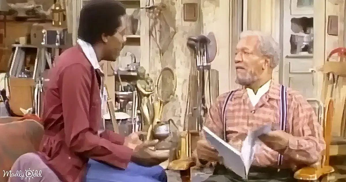 An older man in a plaid shirt holds papers while conversing with a younger man in a red shirt in a cluttered room.