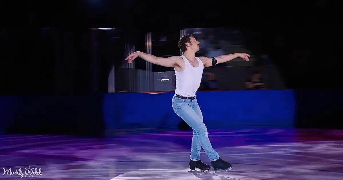 A figure skater in a white tank top and jeans performs a graceful pose on ice.