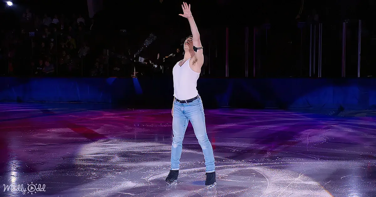 A man in jeans and a white tank top skates on ice with his arm raised, surrounded by a dark audience and colorful lighting.