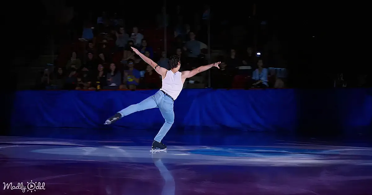 A skater in a white tank top and jeans performs a graceful pose on an ice rink.