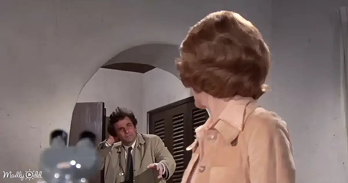 A person in a tan jacket stands facing another person in a room with an arched doorway and wooden shutters.