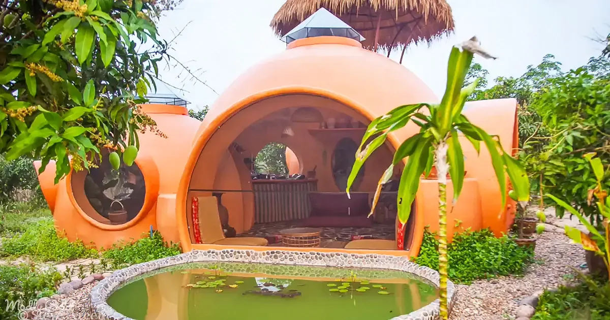 An orange dome-shaped structure with a thatched roof sits amidst lush greenery, featuring a small pond in front.