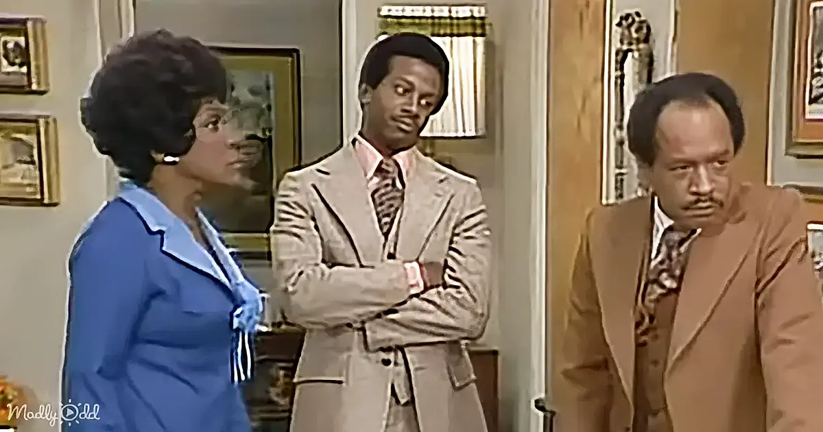 A woman in a blue dress stands beside a man in a tan suit, while another man in a brown suit looks down.
