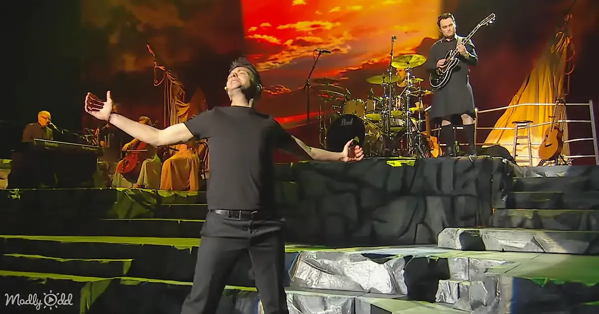 A man in black shirt and pants sings passionately on stage with a band and a fiery background.