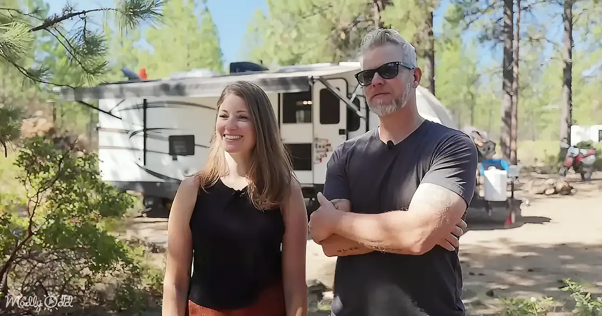 A man and woman stand in front of a white RV in a forested area, with the woman smiling and the man with arms crossed.