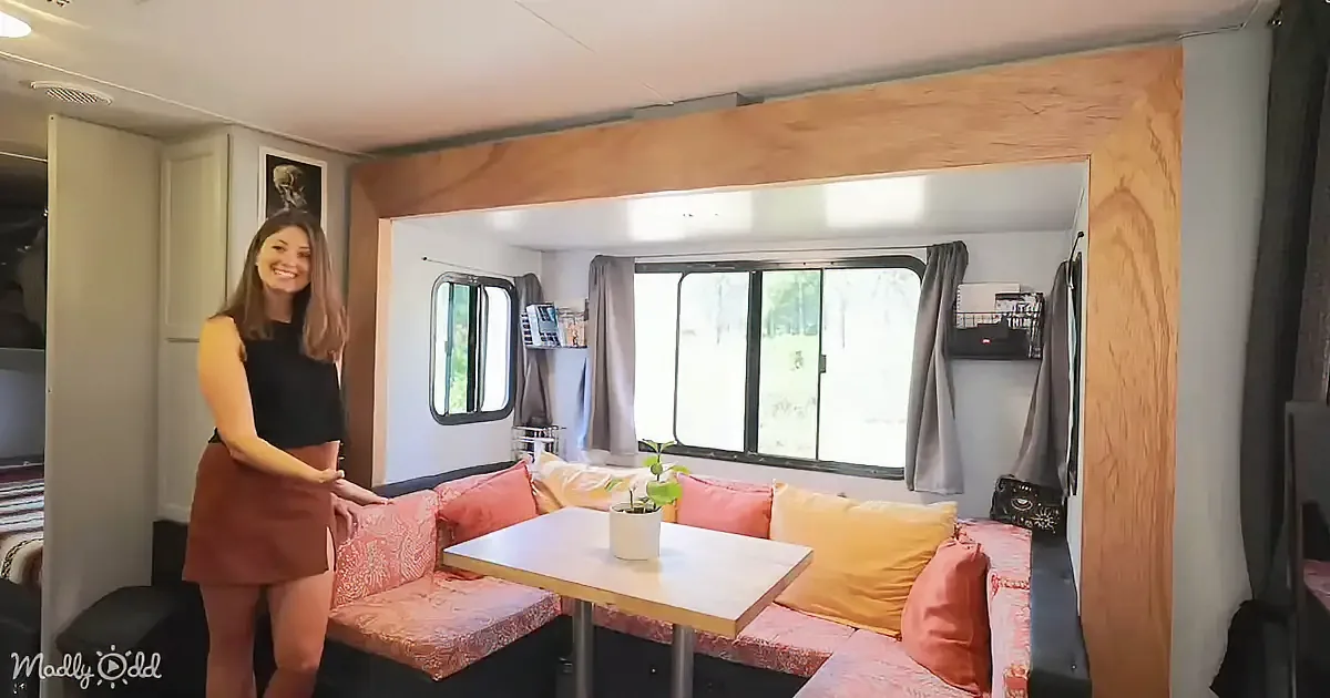 A woman stands in a cozy, compact RV interior with a sofa, table, and large window.