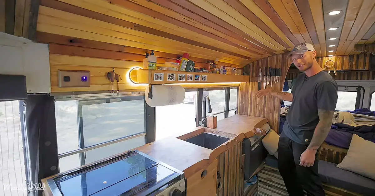 A man stands in a cozy, rustic camper kitchen with wooden walls, a sink, and a stove.