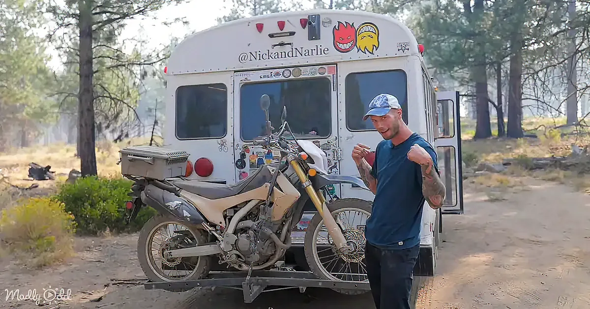 A man stands next to a white bus with a motorcycle on a trailer, surrounded by a forested area.