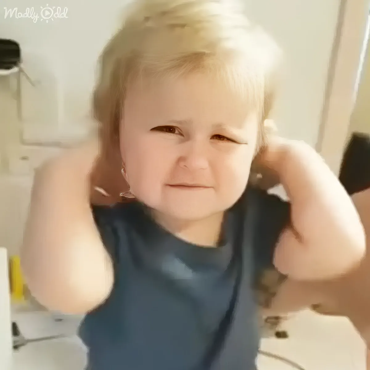 A baby with blonde hair, wearing a dark blue shirt, covers their ears with their hands.