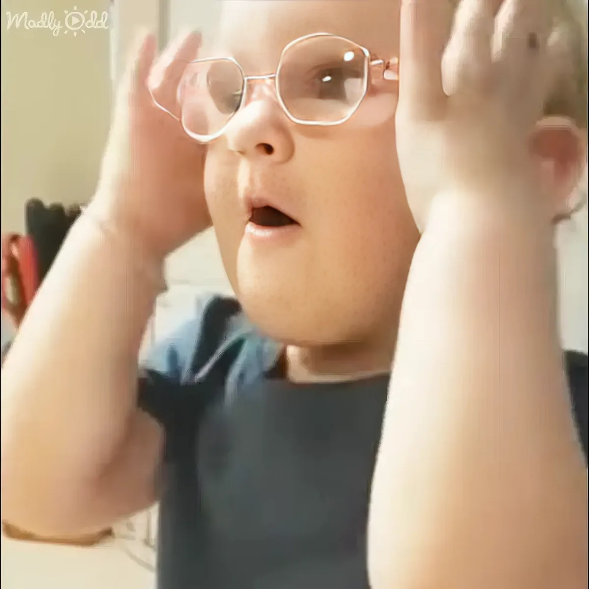 A child wearing glasses and a dark shirt raises both hands near their face.