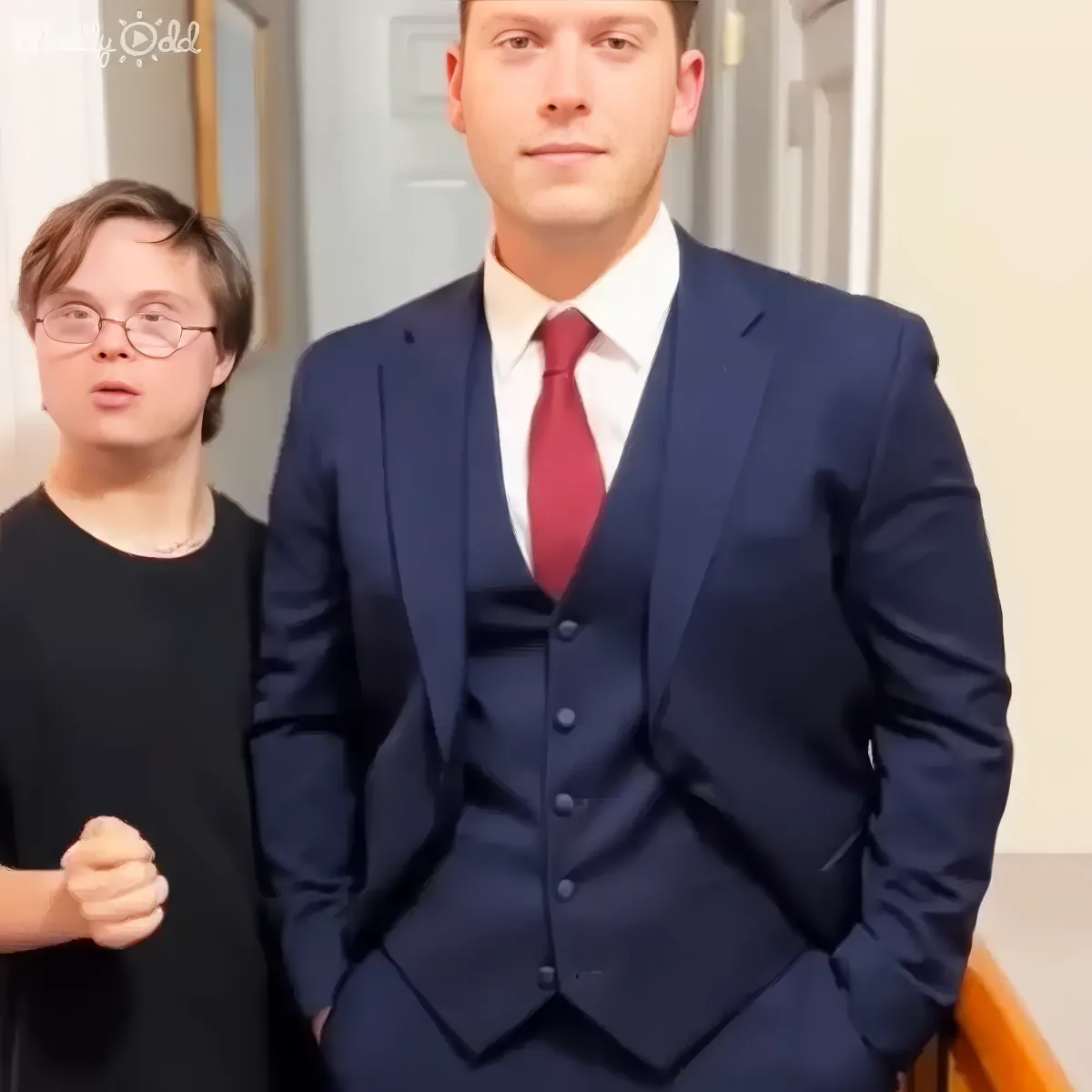 A man in a navy suit with a red tie stands in a hallway, while another person in a black shirt gestures beside him.