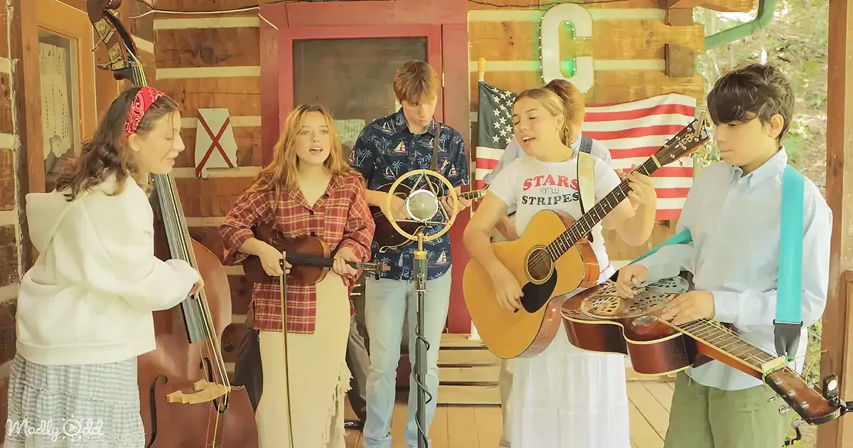 A group of five young musicians perform together, playing instruments and singing in a rustic setting.