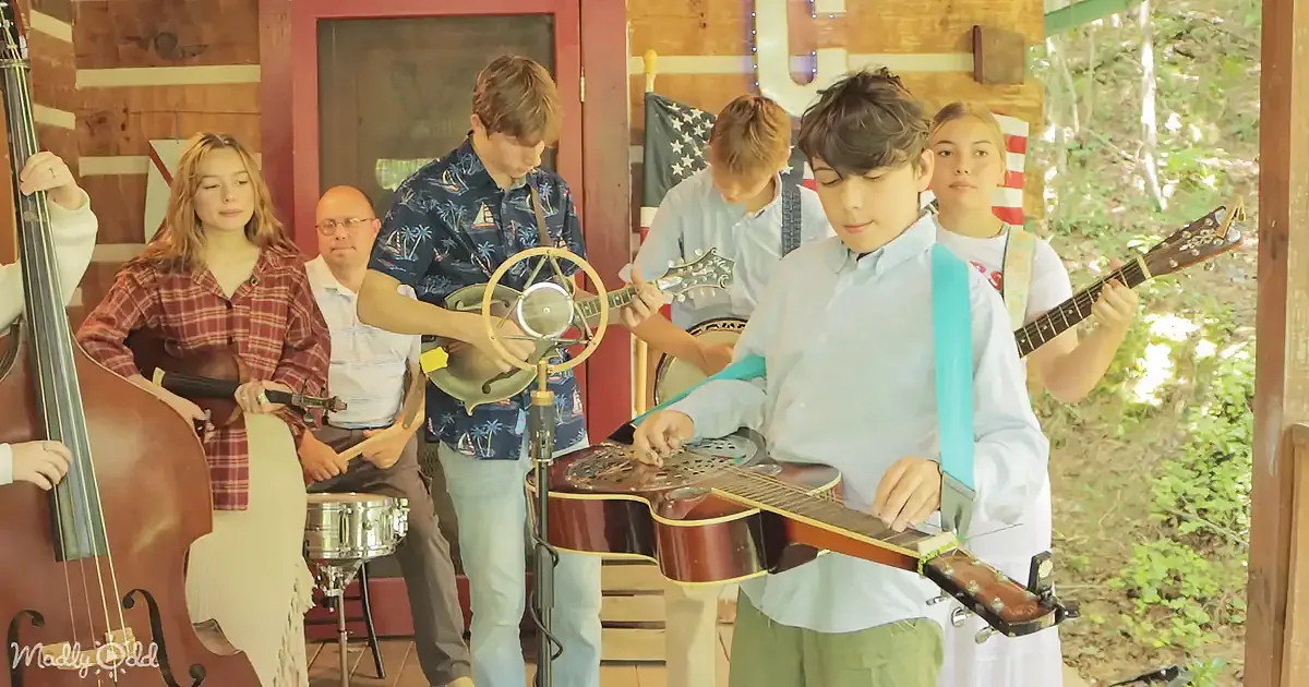 A young boy plays a resonator guitar in front of a group of musicians and an American flag.