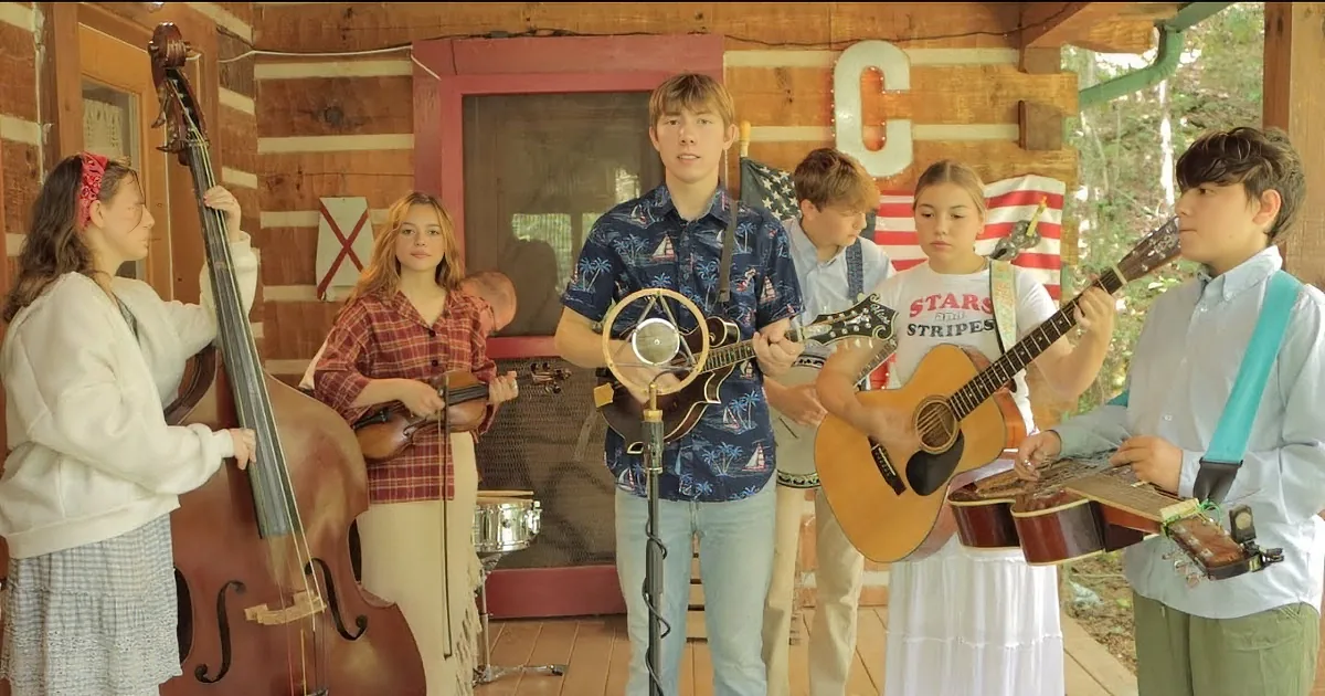 A young man in a blue shirt sings into a microphone while surrounded by musicians playing various instruments.