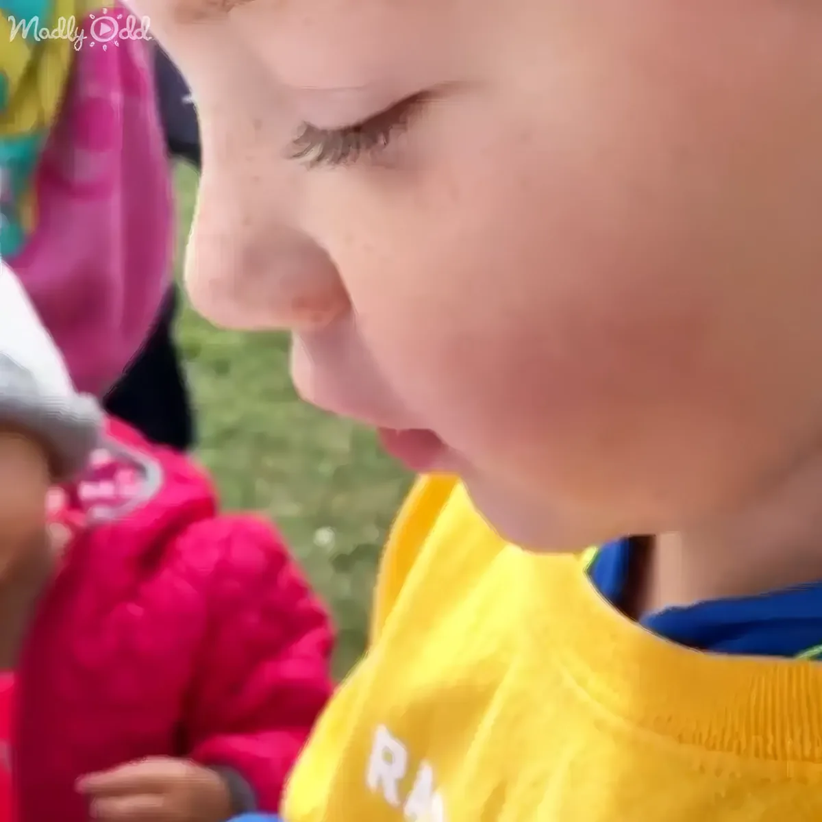 A child in a yellow shirt looks down, with another child in a pink jacket partially visible in the background.