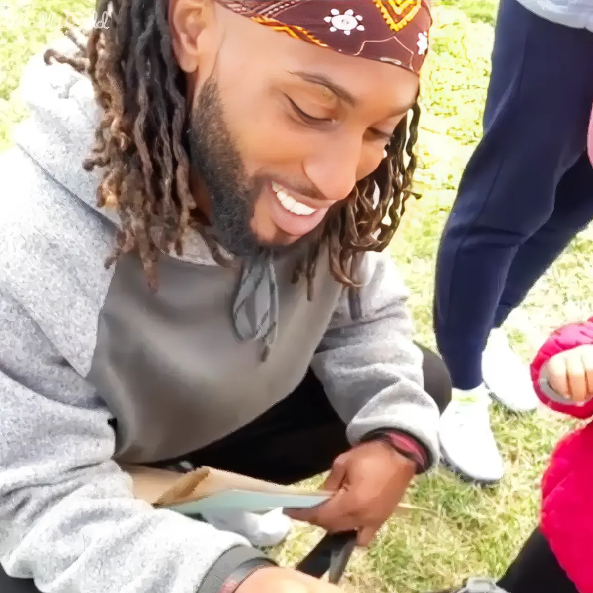 A man with dreadlocks and a bandana smiles while interacting with a child on grass.