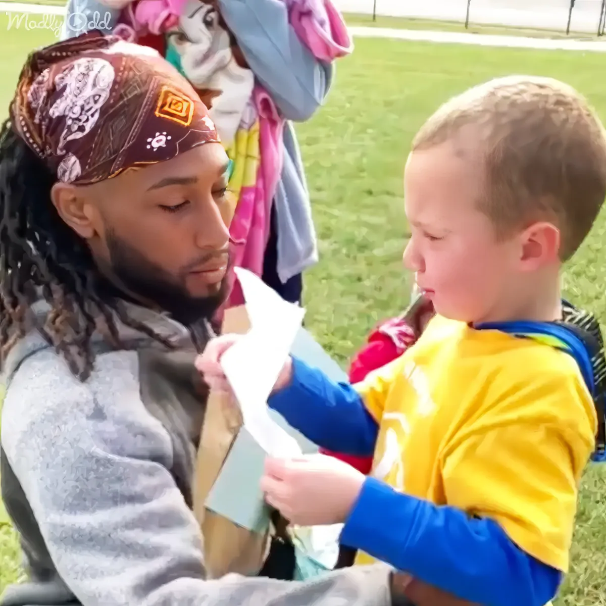 A young boy in a yellow shirt hands a piece of paper to a man with dreadlocks and a bandana in a grassy field.
