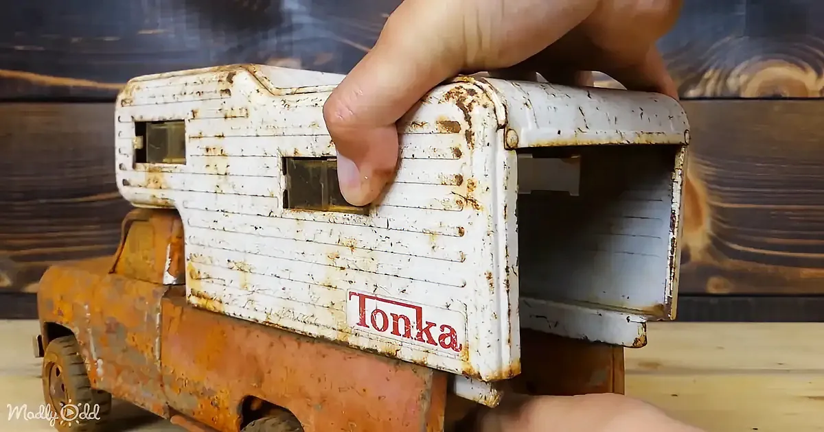 A hand holds a rusty, white Tonka truck with a camper on top, placed on a wooden surface.