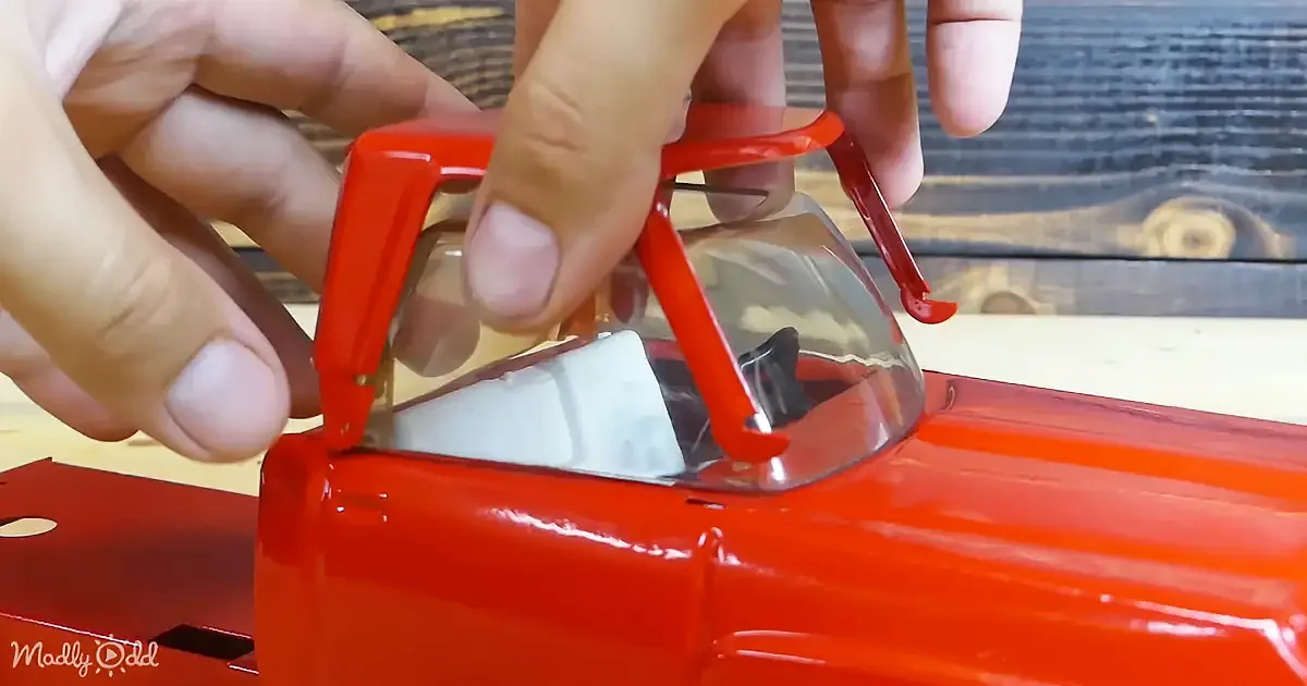 Hands hold a red plastic tool to remove a clear plastic part from a red toy car.