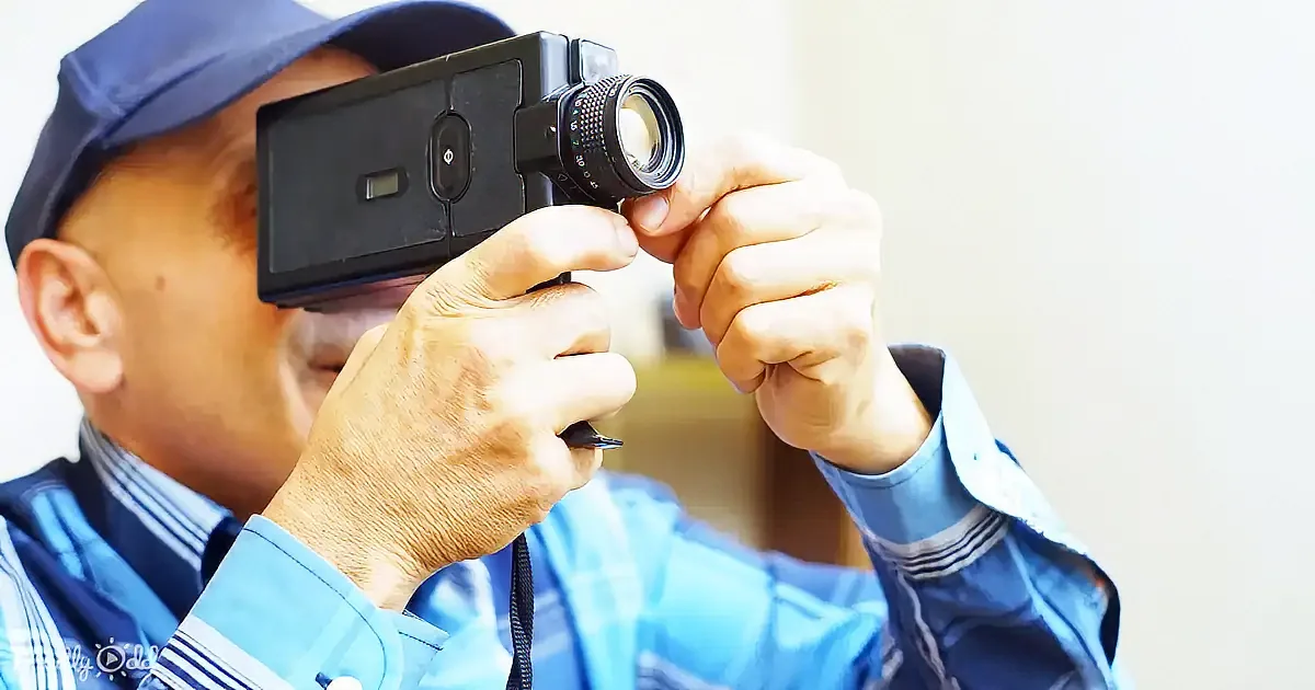 A man in a blue shirt and cap holds a vintage camera, focusing on the lens.
