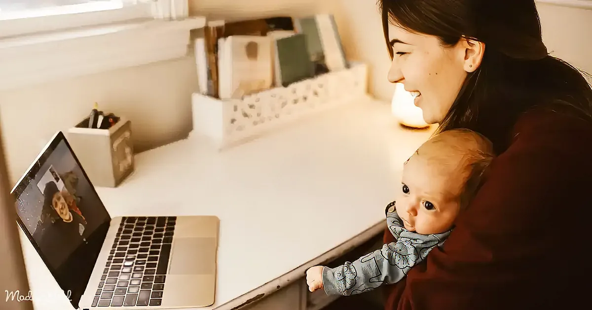 A woman holds a baby while smiling at a laptop screen, possibly during a video call.