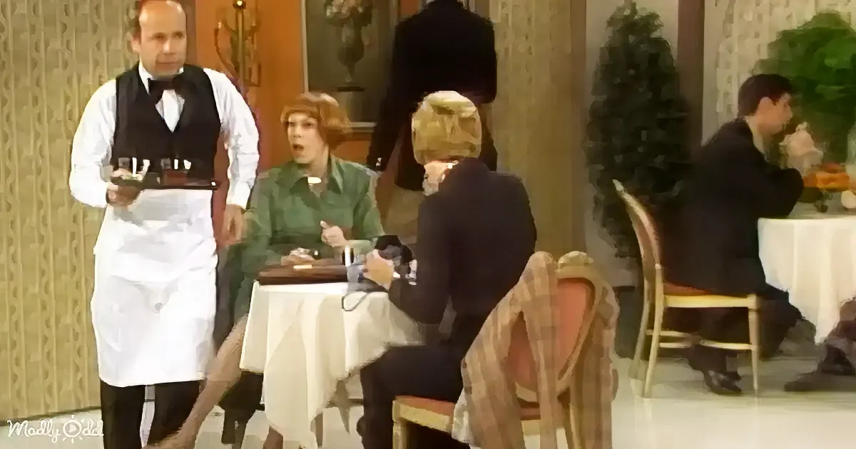 A waiter in a white apron and black vest stands near a table where a woman in a green dress sits, appearing surprised.