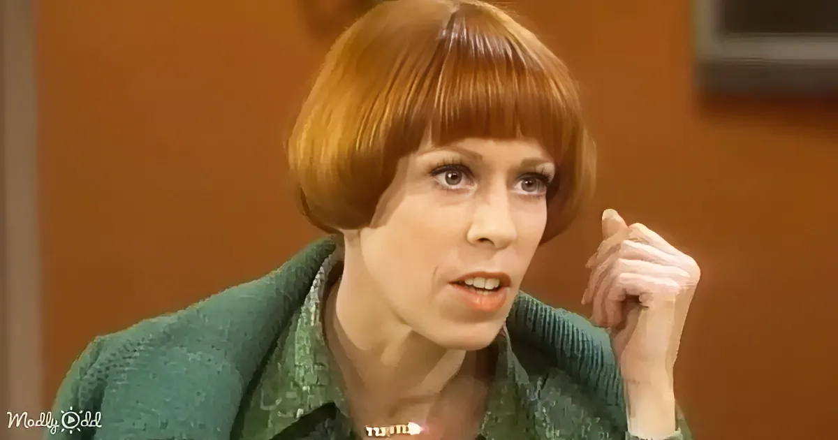 A woman with short red hair and a green sweater is speaking, holding her hand near her face.
