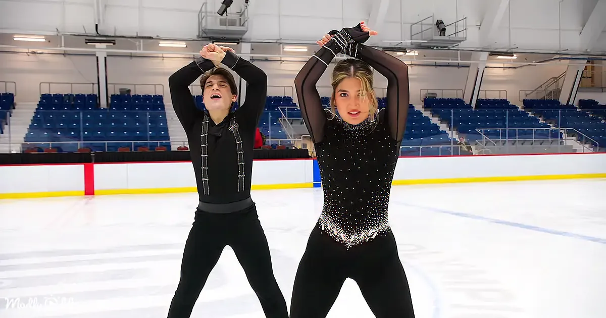 A couple in black figure skating attire stands on an ice rink, arms raised, in a synchronized pose.