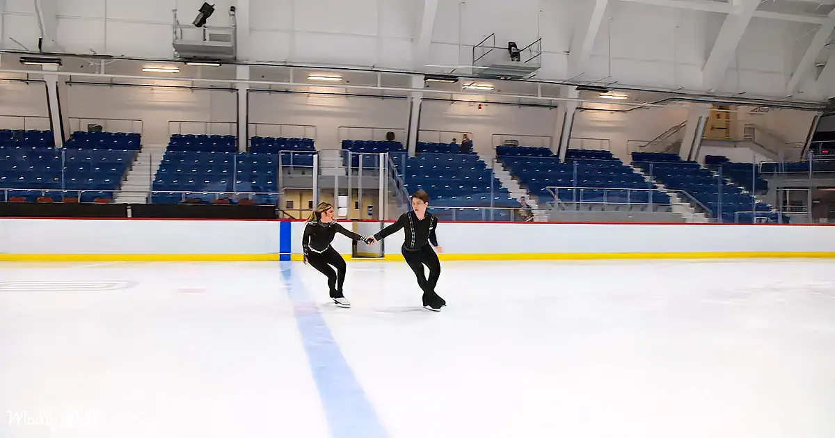 Two figure skaters in black outfits skate hand-in-hand on an indoor ice rink with empty blue seats in the background.