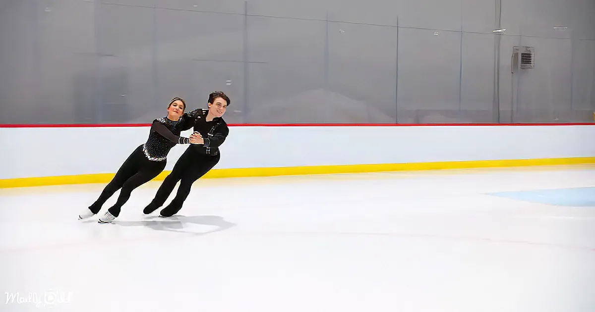 Two figure skaters in black outfits glide on ice, holding hands, with a red and yellow barrier in the background.