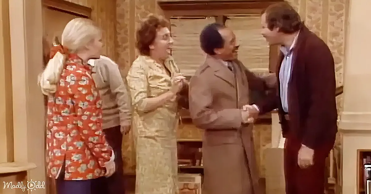 A man in a brown suit shakes hands with another man in a dark suit while two women in floral and yellow dresses watch.