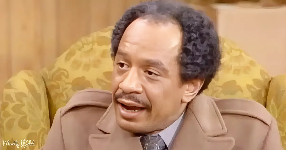 A man with curly hair, wearing a beige suit, sits on a yellow floral-patterned couch, smiling.
