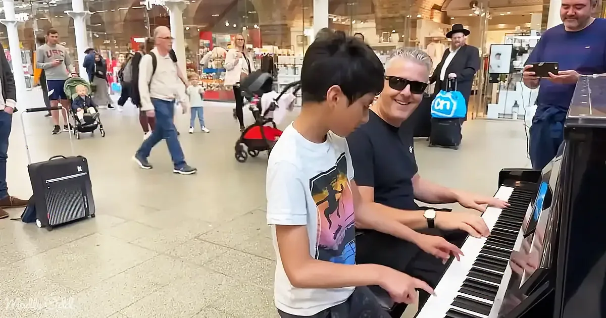 A boy and an adult play piano in a busy airport terminal with people walking by.
