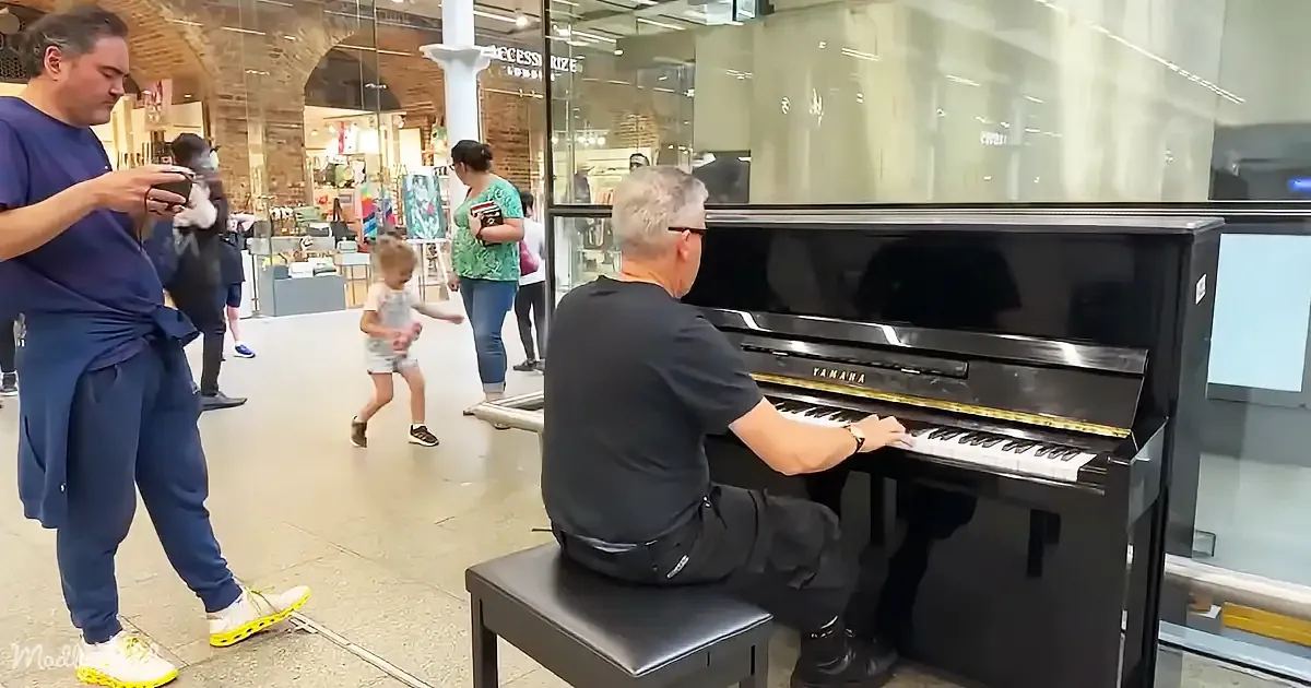 A man in a black shirt plays a Yamaha piano in a shopping mall, while a child dances nearby and others watch.