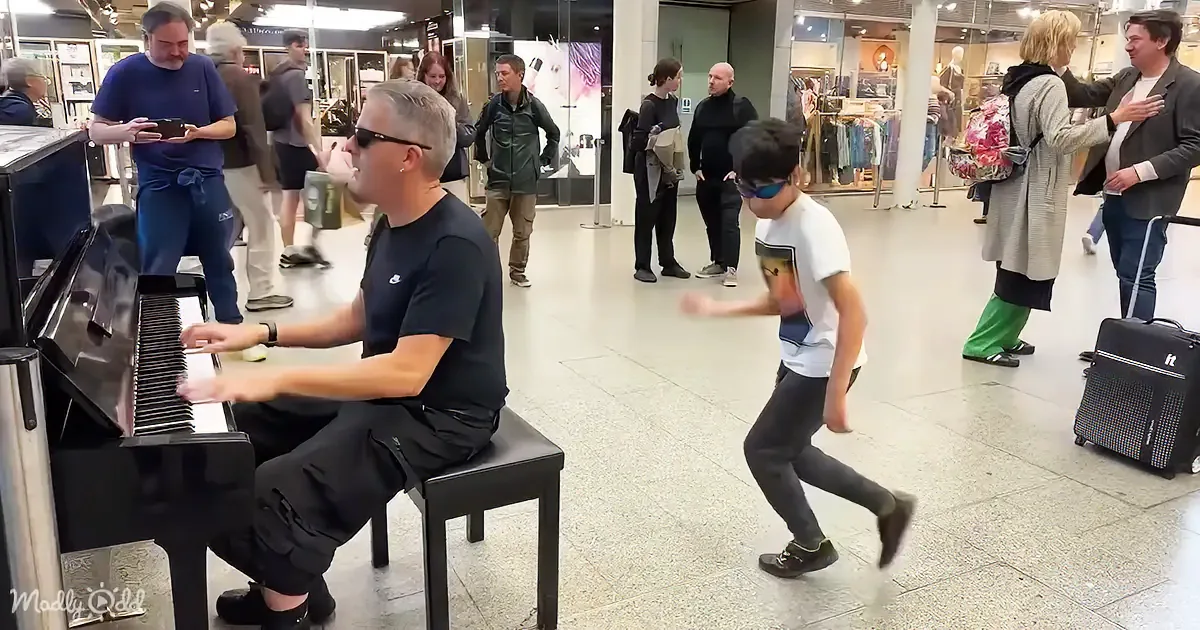 A man plays piano while a boy dances nearby in a busy shopping mall.