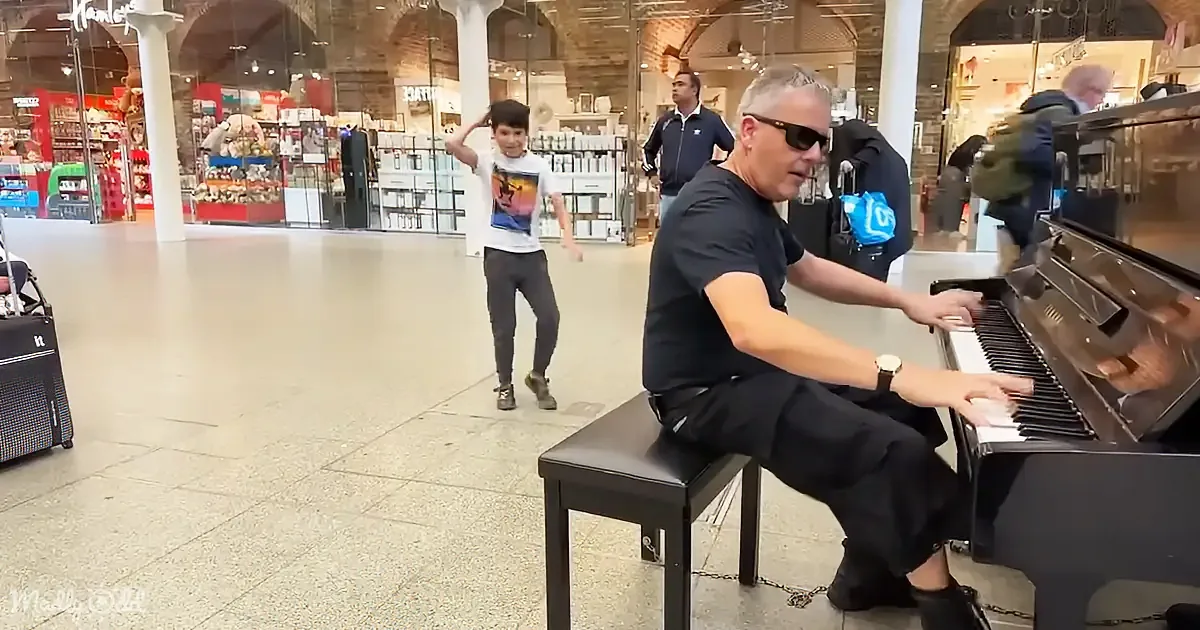 A man plays piano while a boy dances in a bustling shopping mall.
