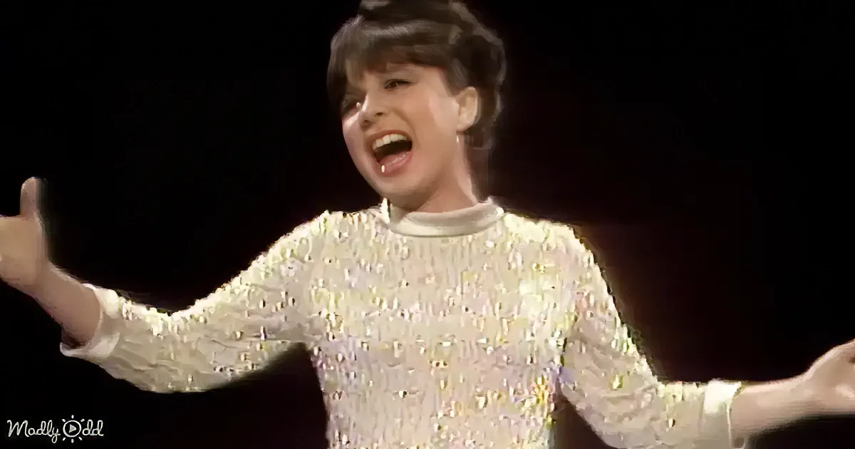 A person in a sparkly, long-sleeved outfit sings passionately with arms outstretched.
