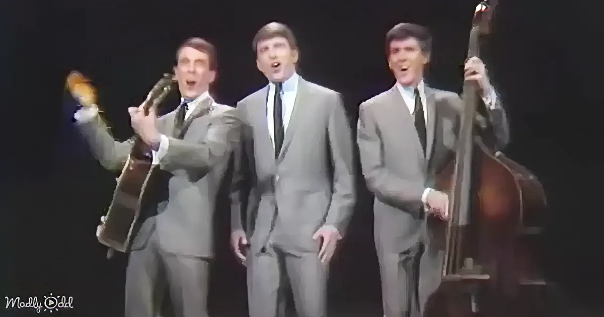Three men in suits sing and play instruments on stage, one holding a guitar, another a double bass.